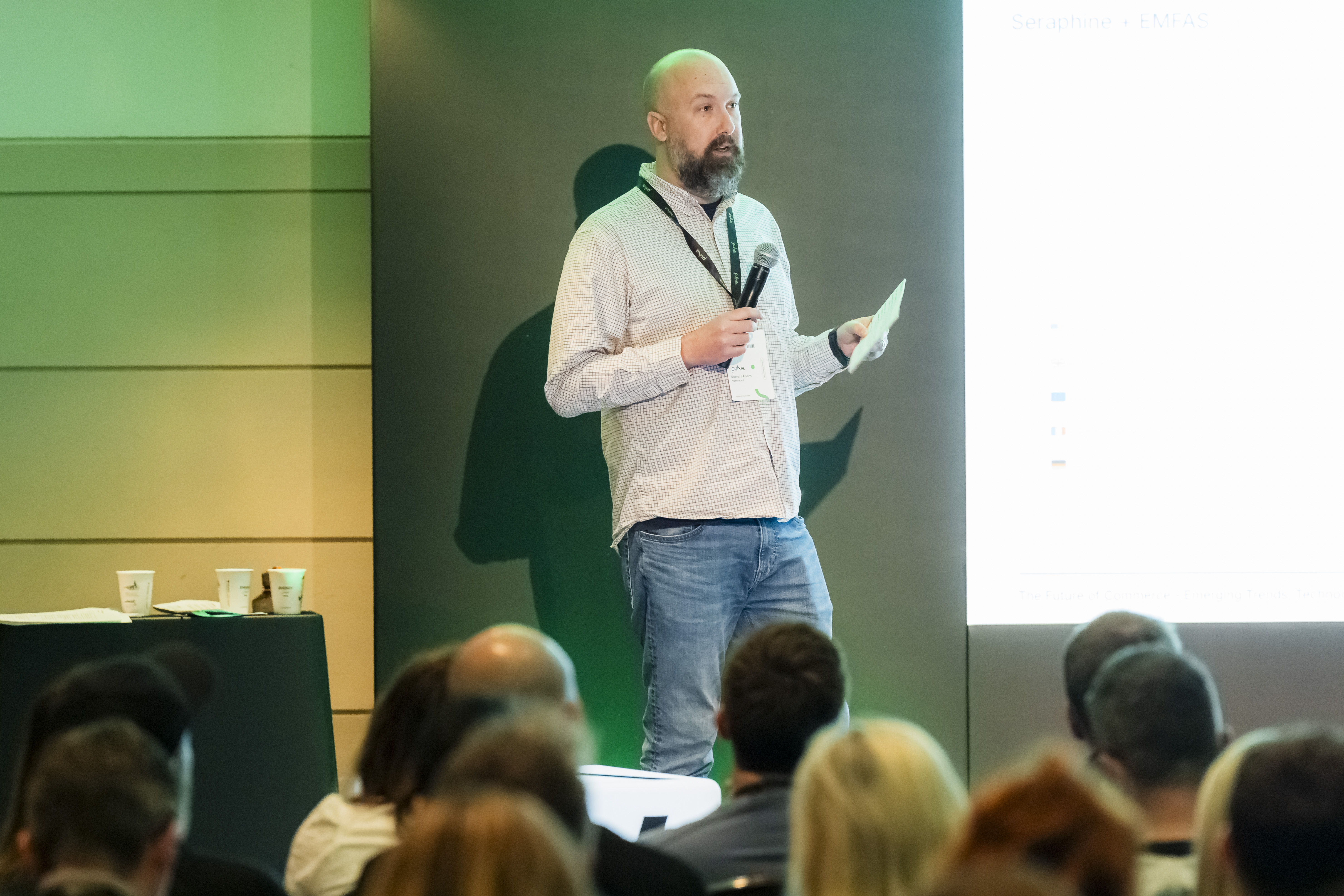 Barrett Ahern delivering a keynote at Pulse eCommerce Summit 2025, speaking on stage to a full conference audience with presentation slides visible on a large screen behind him